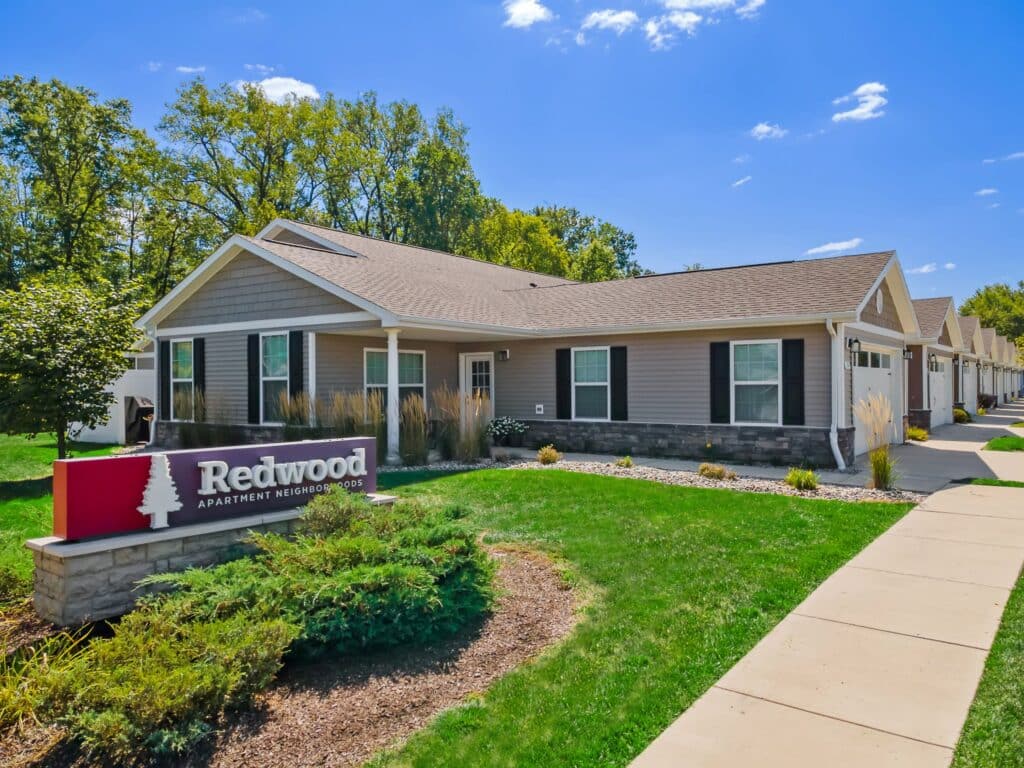 Single-story apartment building with gray siding and stone accents, surrounded by green lawn and landscaping, with a sign that reads "Redwood Apartment Neighborhoods.