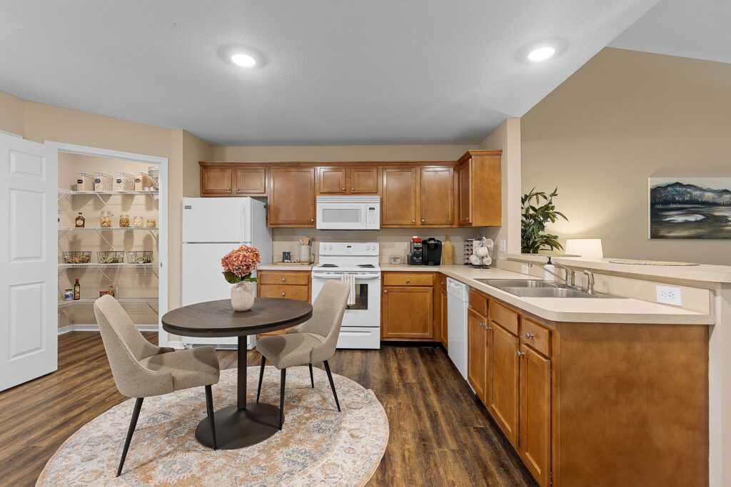 A kitchen with wooden cabinets, white appliances, a round dining table with two chairs, and an open pantry stocked with food items.