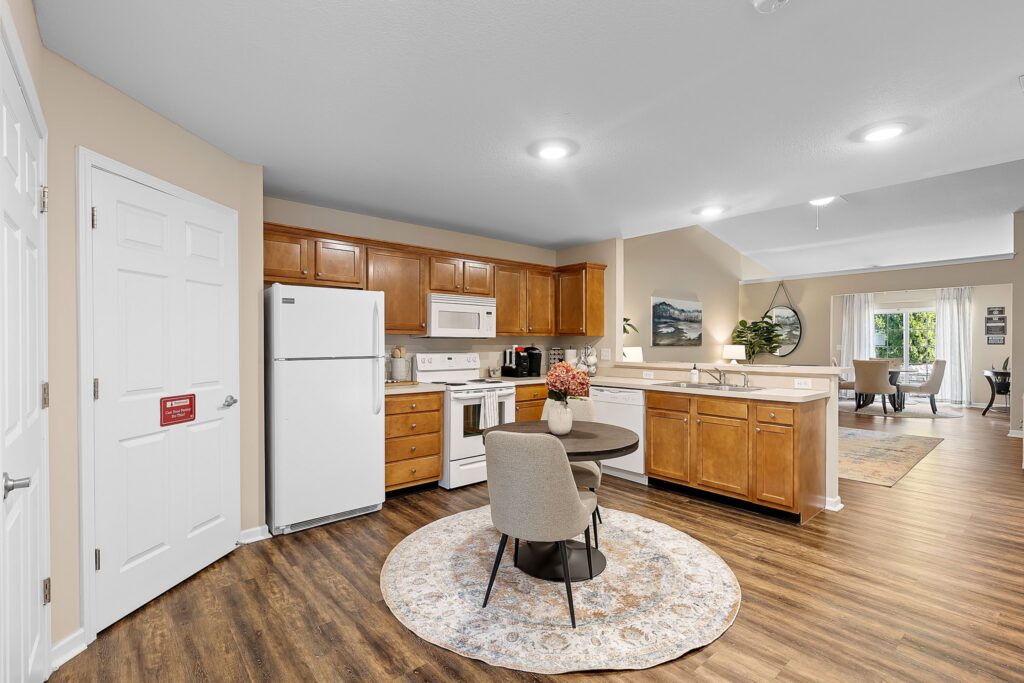 Modern kitchen with wood cabinets, white appliances, and round dining table on a patterned rug; open layout connects to a living area with large windows.