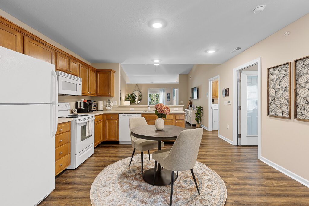 A modern kitchen with wooden cabinets, white appliances, and a round dining table with four chairs on a patterned rug, featuring wall art and neutral décor.