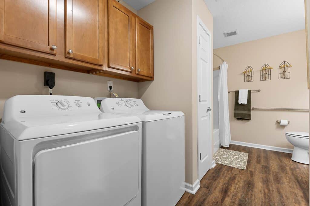 Laundry room with a white washer and dryer, wooden cabinets above, and an adjacent bathroom area featuring a toilet, towel rack, and shower with a curtain.