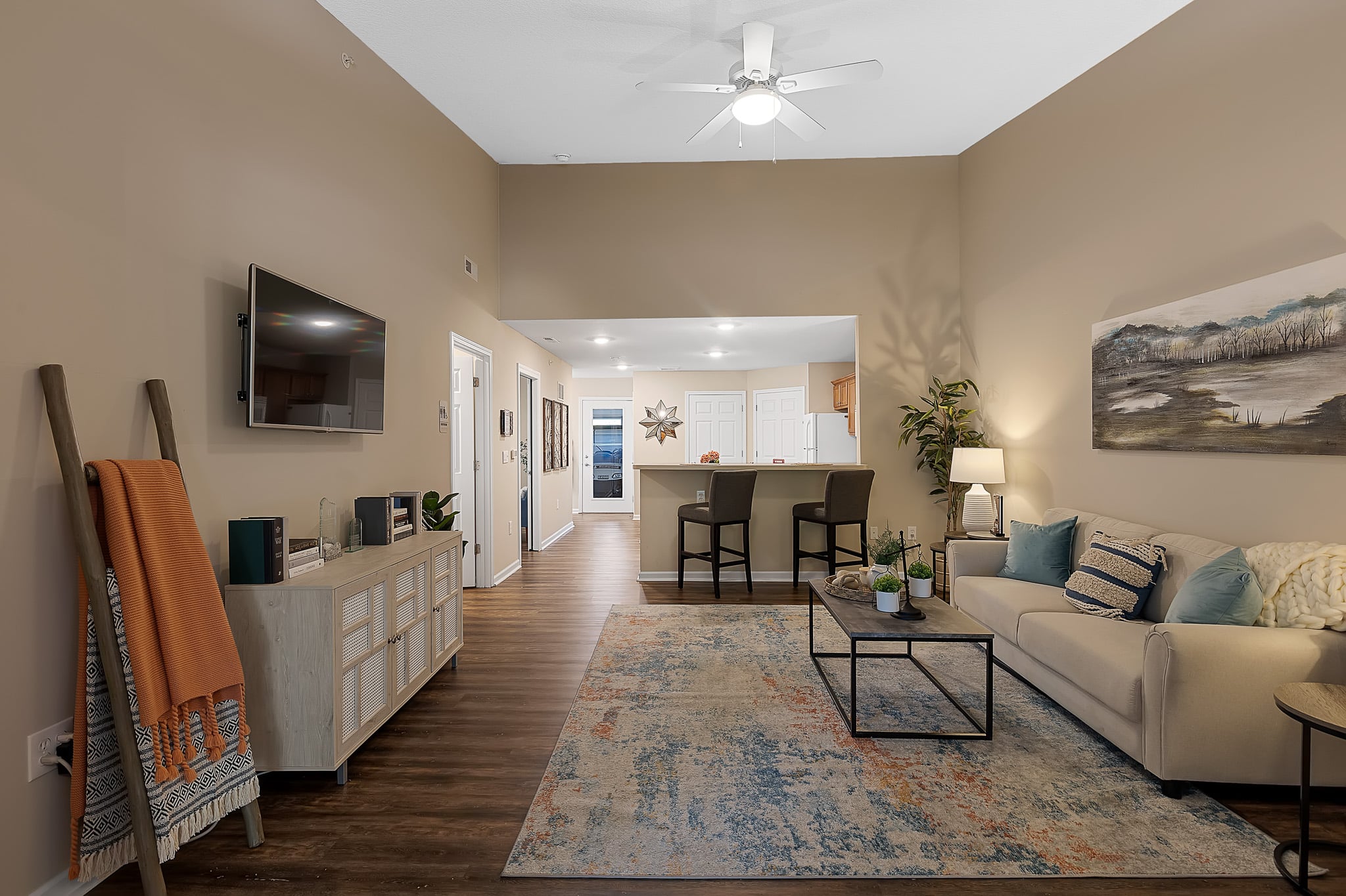 Modern living room with a beige sofa, patterned rug, wall-mounted TV, breakfast bar, and neutral decor leading into a kitchen area with white cabinets.