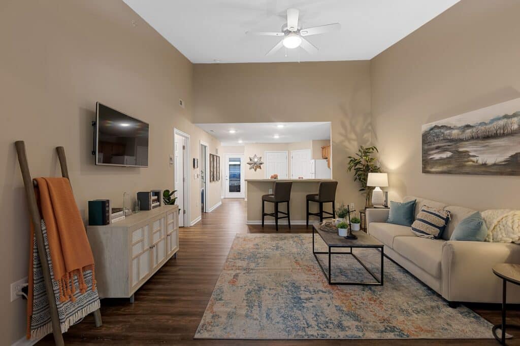A modern living room with a beige sofa, patterned rug, TV, and bar counter leading to a kitchen; decor includes plants, wall art, and neutral-toned furnishings.