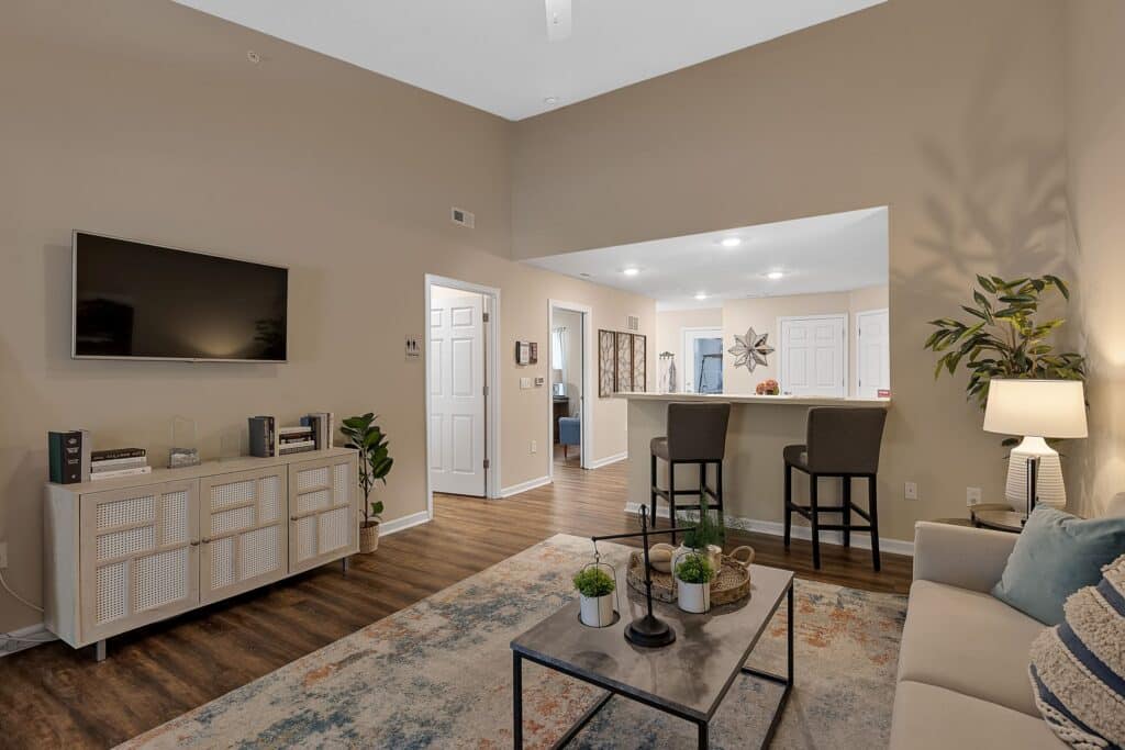 Modern living room with a wall-mounted TV, a sofa, coffee table with plants, a rug, and a kitchen bar area with two chairs in an open floor plan.