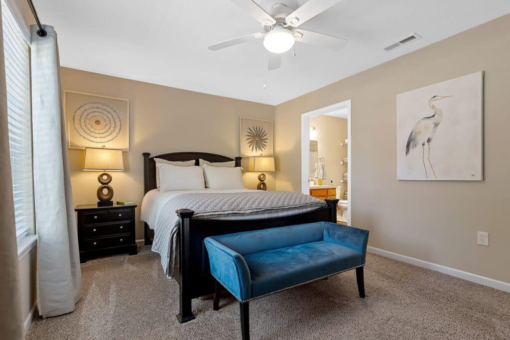 A bedroom with a double bed, two bedside tables with lamps, a blue upholstered bench at the foot of the bed, beige walls, and artwork on the walls.