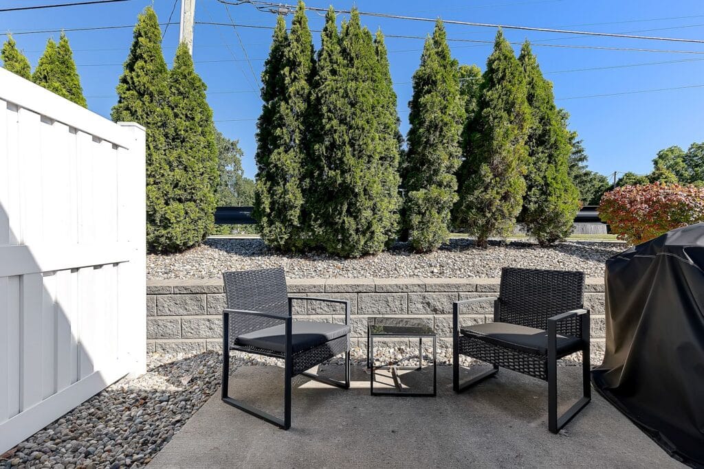 Two black patio chairs and a small glass-top table on a concrete patio, with tall evergreen trees and a white fence in the background.