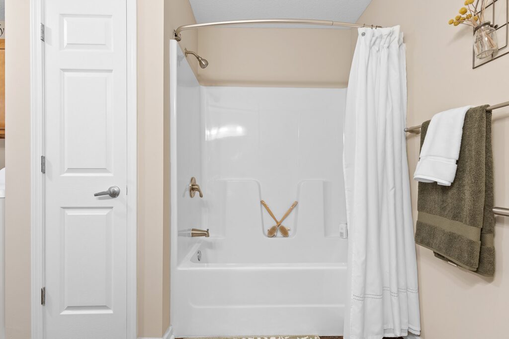 White bathtub with shower curtain partially open, brass fixtures, and two bath brushes inside. Towel rack with folded towels on beige wall beside the tub.