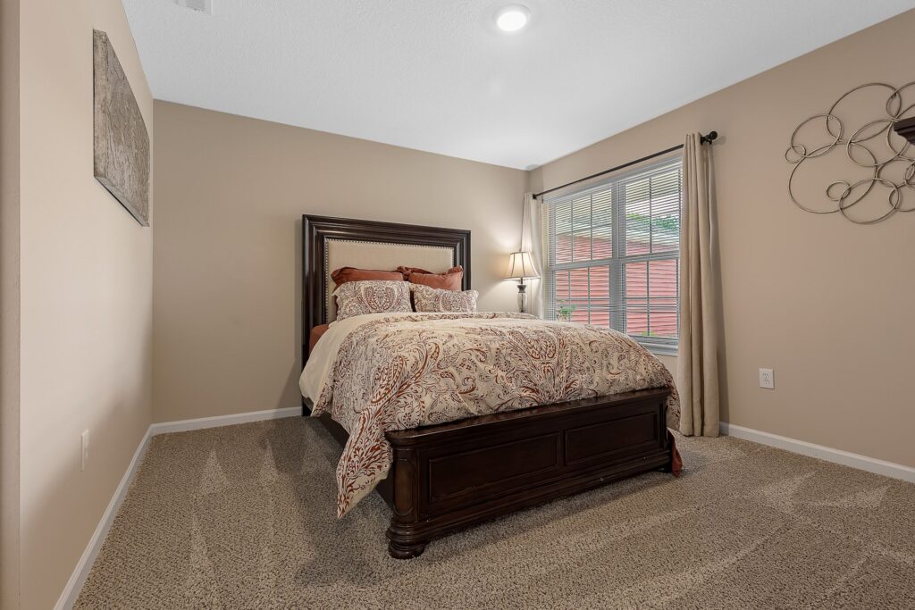 A neatly made bed with patterned bedding is placed against the wall in a beige-carpeted bedroom with tan walls, a window with blinds, and a decorative wall piece.