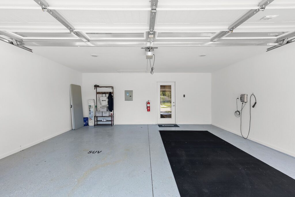 Clean, empty two-car garage with white walls, a storage shelf on the left, wall outlets, a fire extinguisher, a mat, and a door leading outside at the back.