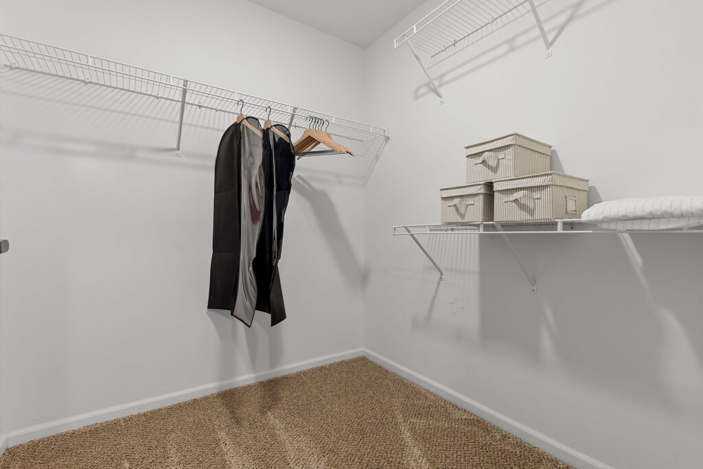 A walk-in closet with wire shelving, a few hanging clothes, three beige storage boxes, and a folded white blanket on carpeted flooring.