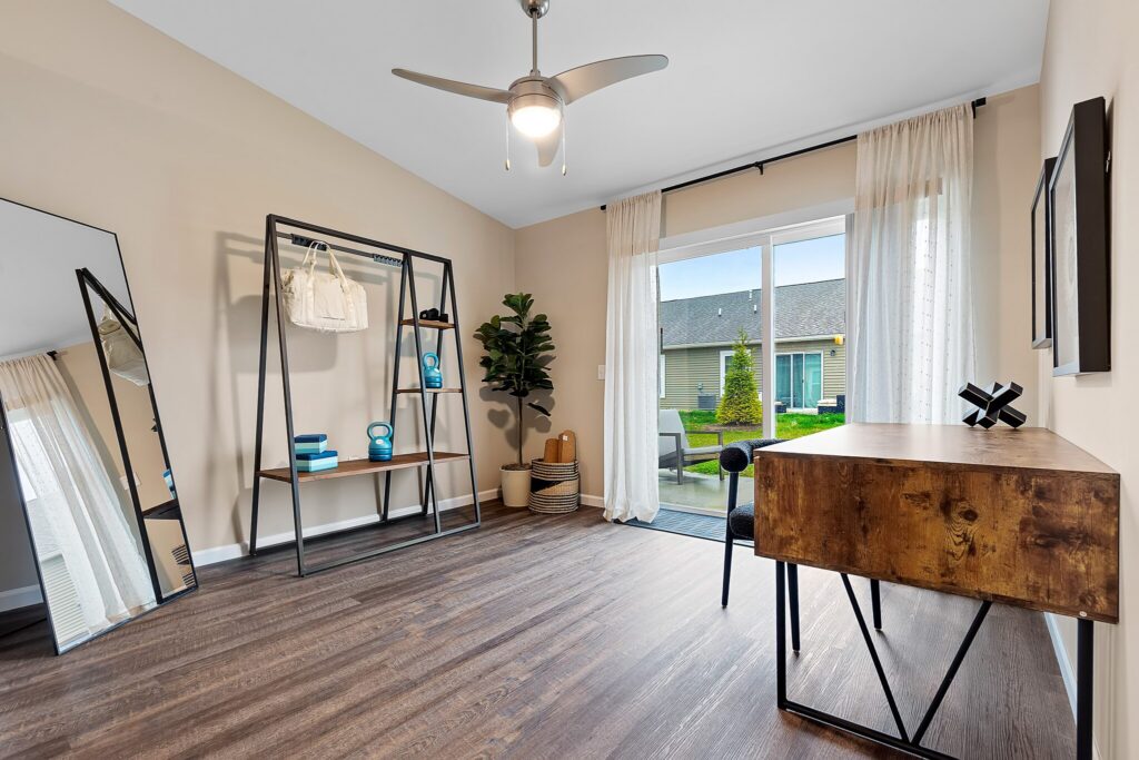 Minimalist home office with a wooden desk, black chair, large mirror, open shelving unit, potted plant, and sliding glass doors opening to a patio and yard.
