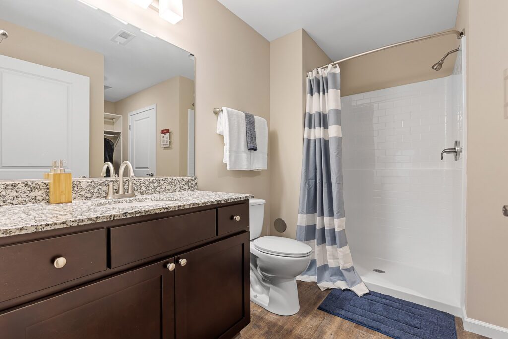 Modern bathroom with granite countertop, dark wood vanity, toilet, striped shower curtain, white tiled shower, towels, and a blue bath mat on wood-style flooring.