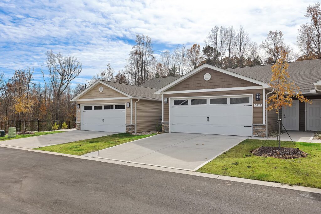 Two attached single-story homes with double garages, concrete driveways, and small lawns, set in a suburban neighborhood with trees in the background.