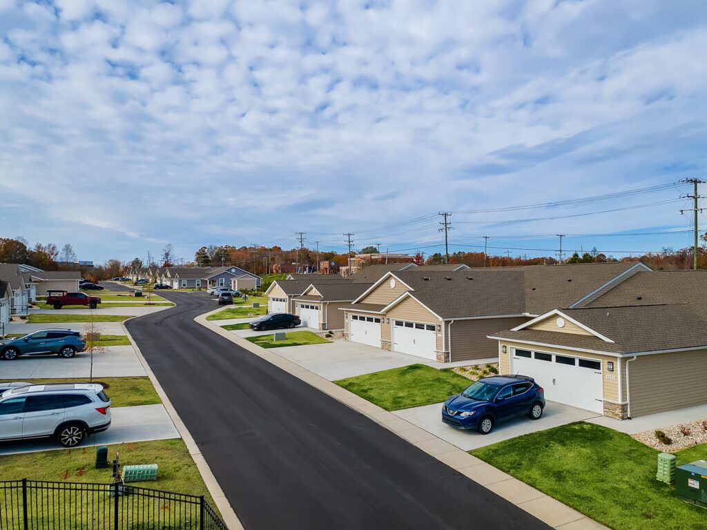 A suburban neighborhood street with single-story houses, driveways, lawns, and several parked cars under a partly cloudy sky.