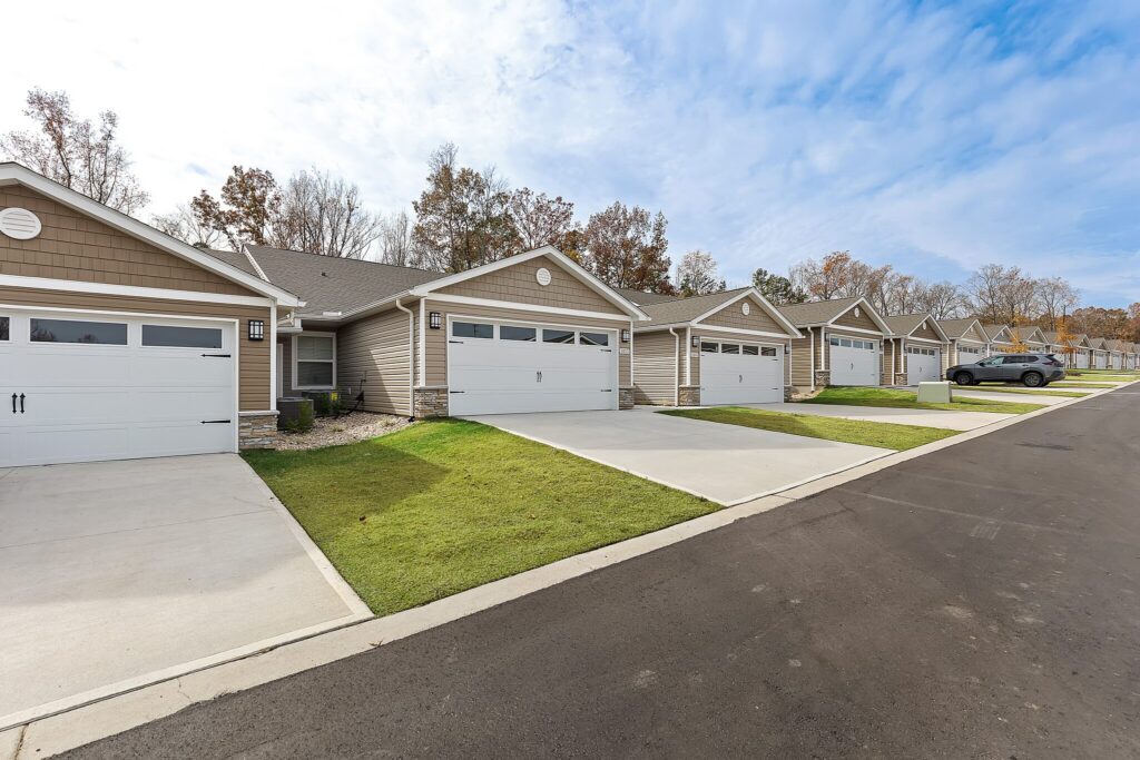 Row of single-story suburban houses with attached garages and driveways, manicured lawns, and a parked car along a paved residential street under a partly cloudy sky.