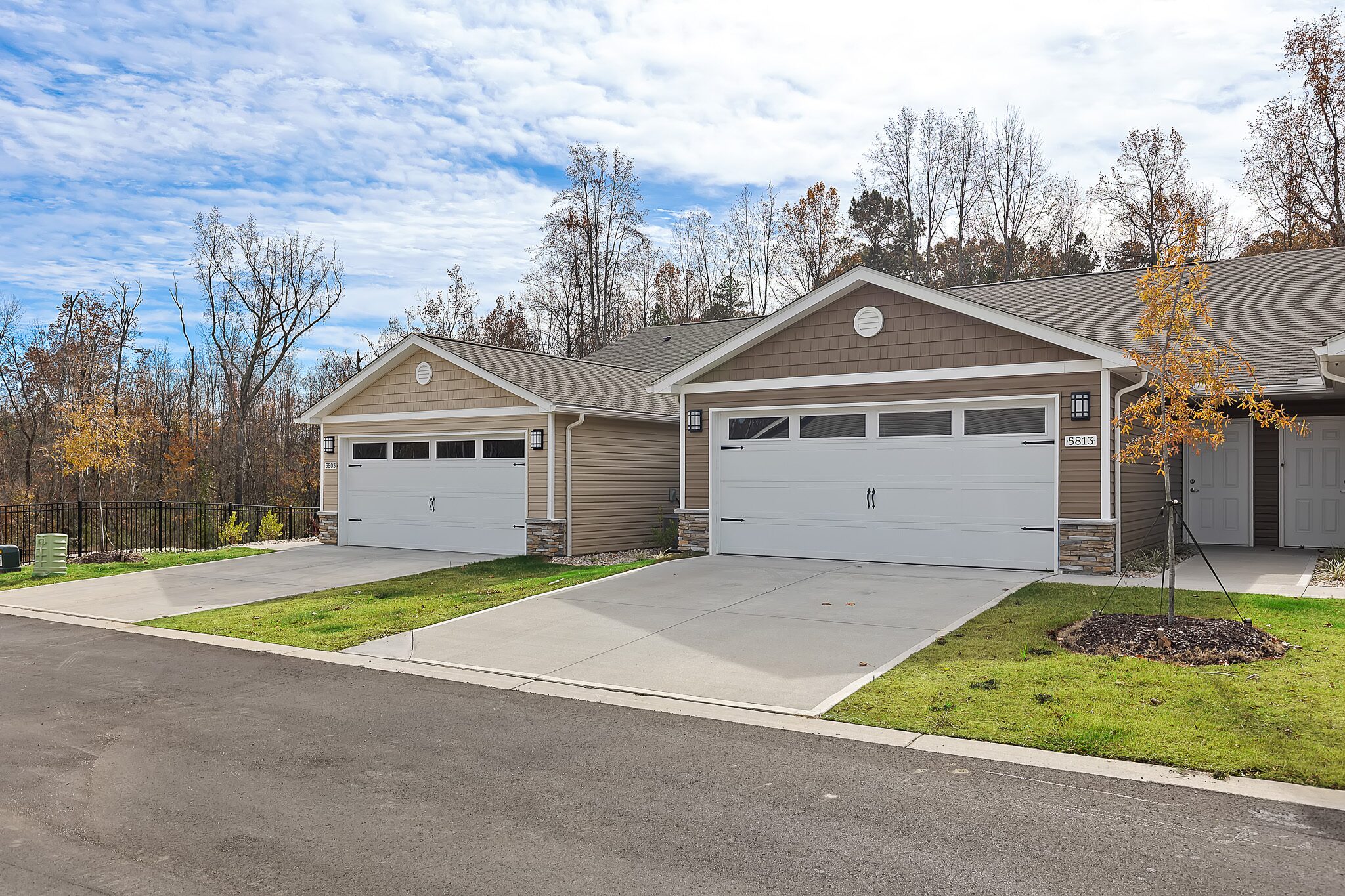 Two attached single-story homes with double garages, concrete driveways, and small lawns, set in a suburban neighborhood with trees in the background.