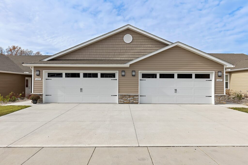 A duplex with tan siding and two white double garage doors, each with windows. The driveway and sidewalks are concrete, with minimal landscaping on the sides.