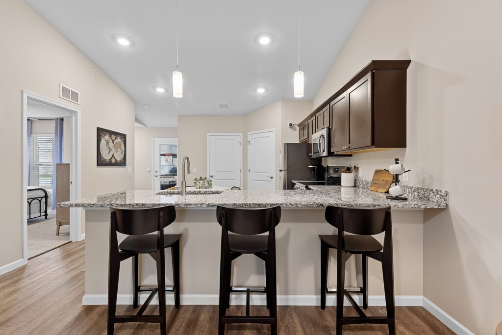 Modern kitchen with granite countertop, three dark wooden barstools, pendant lighting, stainless steel appliances, and beige walls; bedroom visible through open doorway.