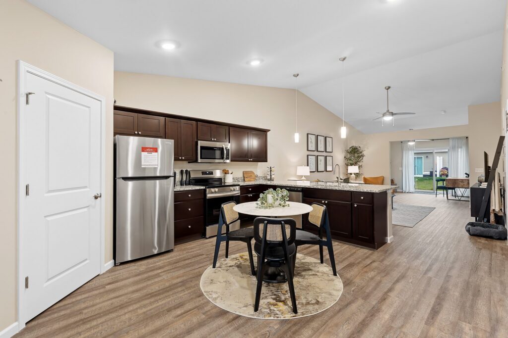Open-concept kitchen and dining area with dark wood cabinets, stainless steel appliances, round dining table, and adjacent living space with large windows.