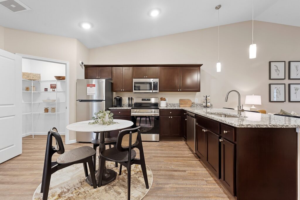 Modern kitchen with dark wood cabinets, stainless steel appliances, granite countertops, a small round dining table with chairs, and an open pantry door on the left.