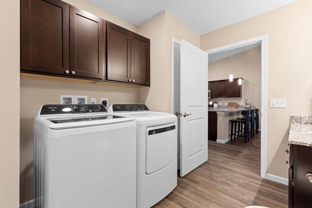 Laundry room with a washing machine and dryer, dark wood cabinets above, beige walls, and a door leading to a kitchen area with similar cabinetry.