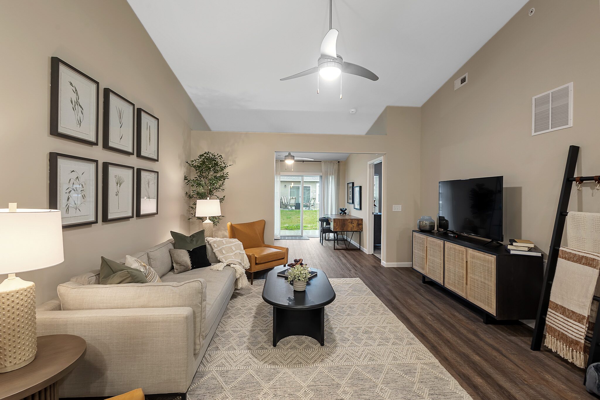 Modern living room with neutral tones, a sofa, armchairs, wall art, TV console, coffee table, and large window opening to a sunroom with outdoor view.