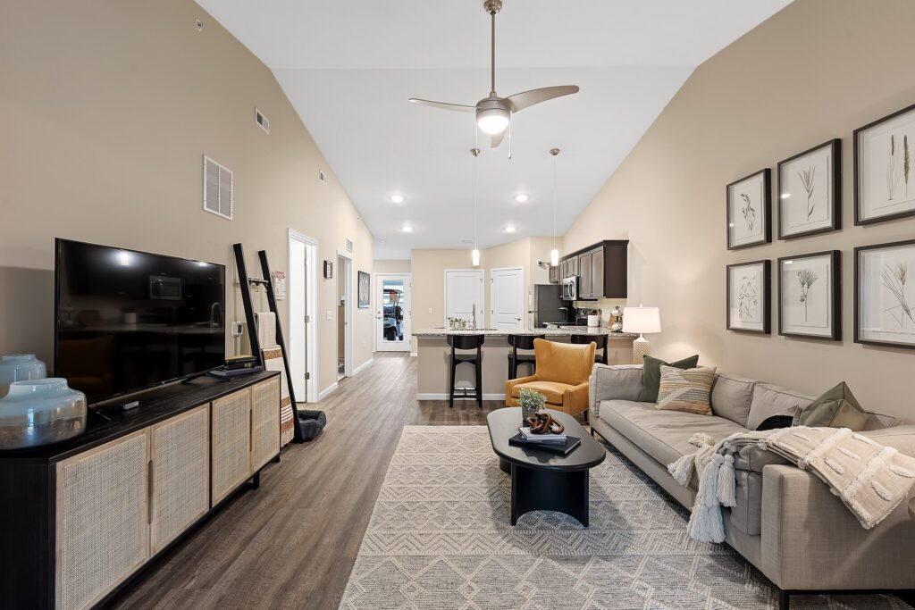 Modern open-concept living room and kitchen with neutral tones, a sofa, wall art, TV, bar stools, and pendant lighting.