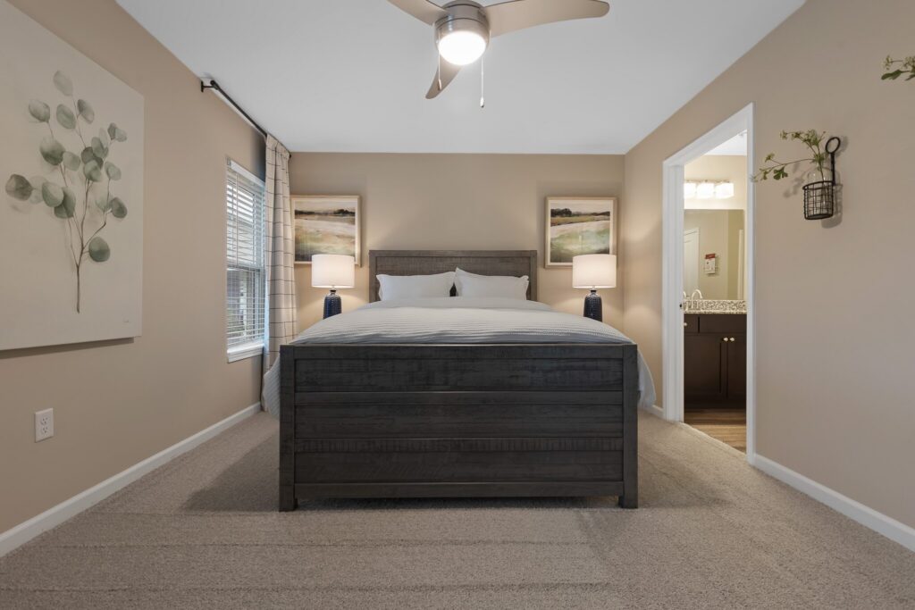 A modern bedroom with a dark wood bed, two nightstands with lamps, neutral walls, carpeted floor, and an open doorway to a bathroom.