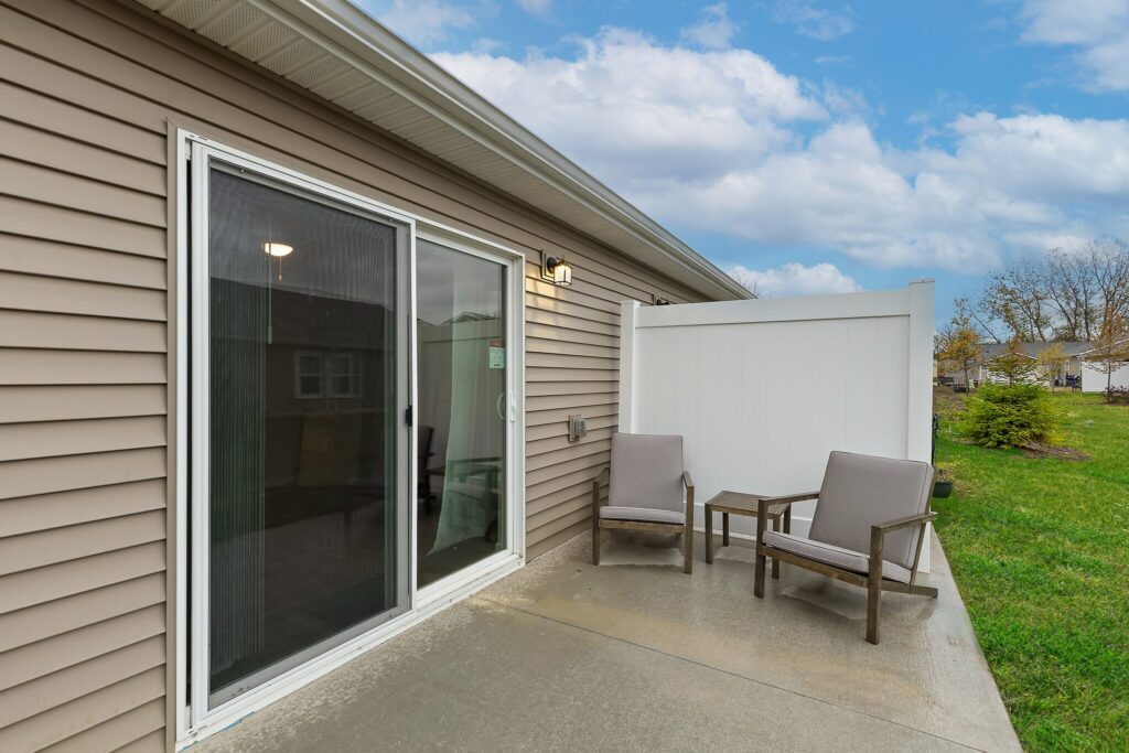 Concrete patio with two cushioned chairs and a small table next to a sliding glass door, bordered by tan siding and a white privacy fence, with grass and trees in the background.