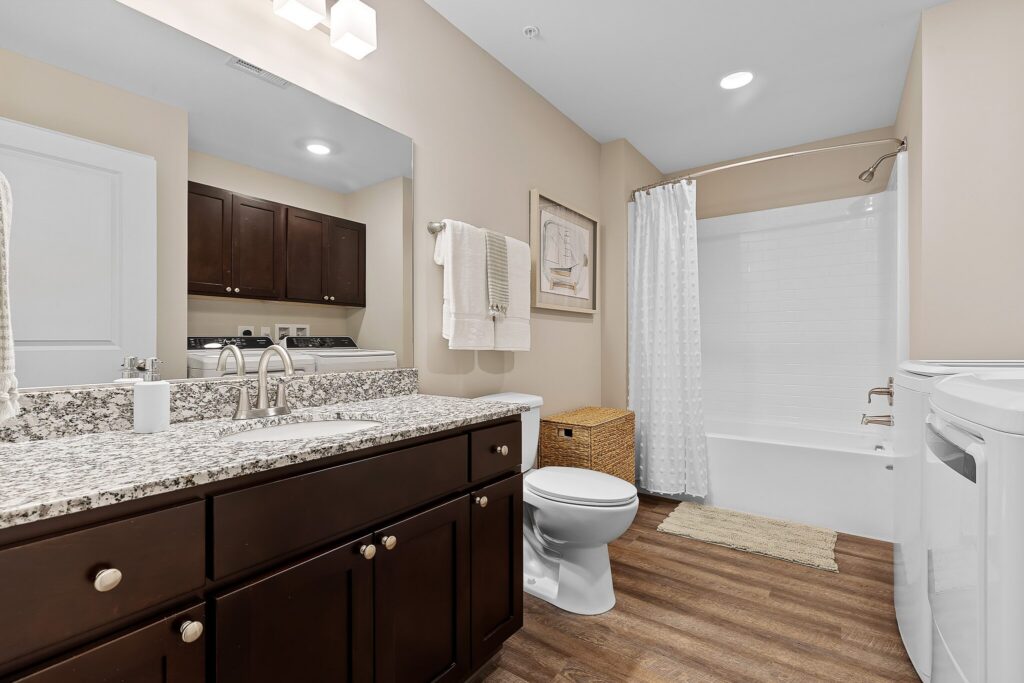 Modern bathroom with double sink vanity, granite countertop, dark wood cabinets, toilet, white shower-tub combo with curtain, wall art, and wood-style flooring.