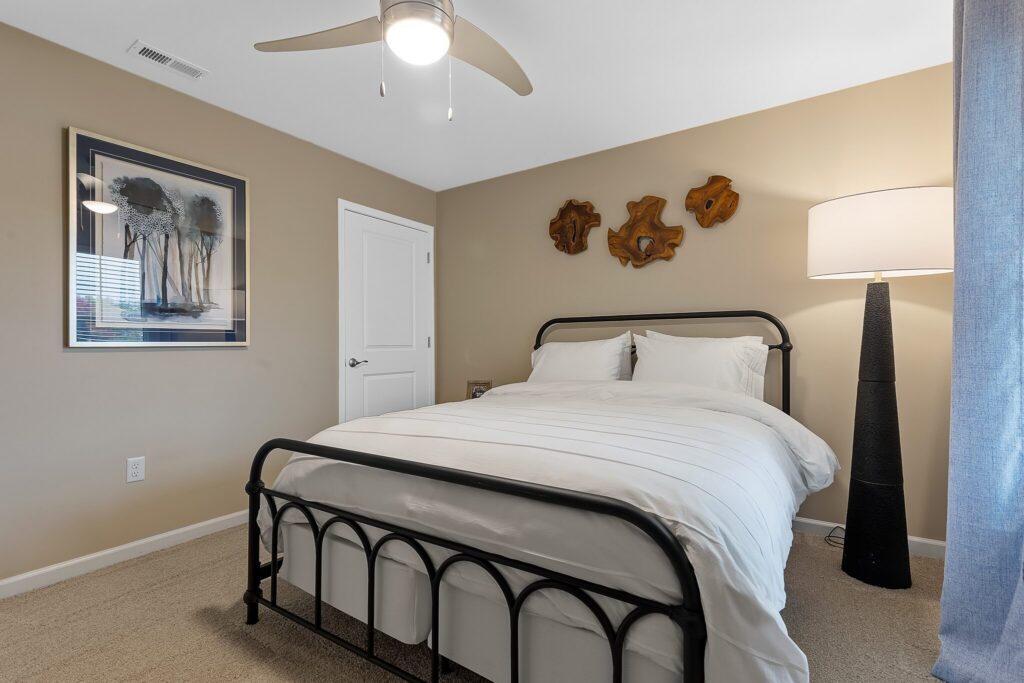Bedroom with a black metal bed frame, white bedding, a floor lamp, wall art, and a ceiling fan in a beige room with carpeted flooring.