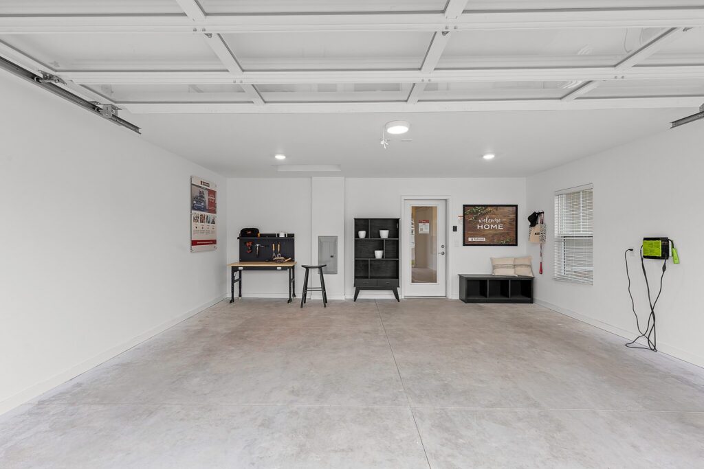 A clean, empty garage with white walls, a workbench, shelves, and an electric vehicle charger mounted on the right wall. Door and windows are visible in the back.