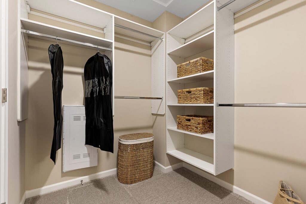 Walk-in closet with hanging rods, white shelves holding wicker baskets, two black garment bags, and a woven laundry hamper on the carpeted floor.