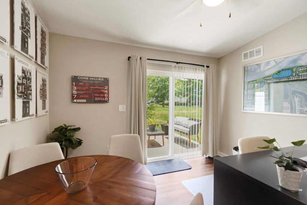 Dining area with a round wooden table, four chairs, wall art, and glass doors opening to a green backyard. Light curtains hang by the doors, and plants are in the corners.
