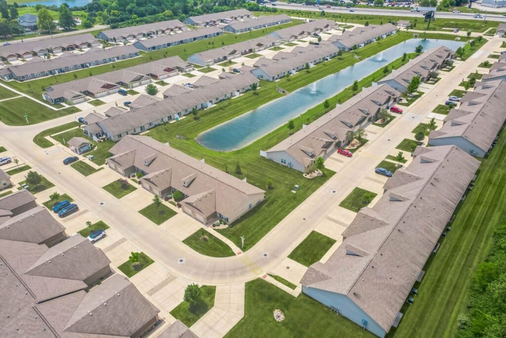 Aerial view of a suburban residential neighborhood with rows of houses, driveways, green lawns, and a central pond.