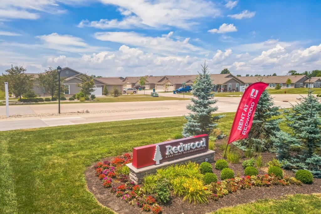 Entrance sign for Redwood Apartments & Townhomes, surrounded by landscaped flowers and bushes, with a red "Rent at Redwood" flag and a suburban neighborhood in the background.
