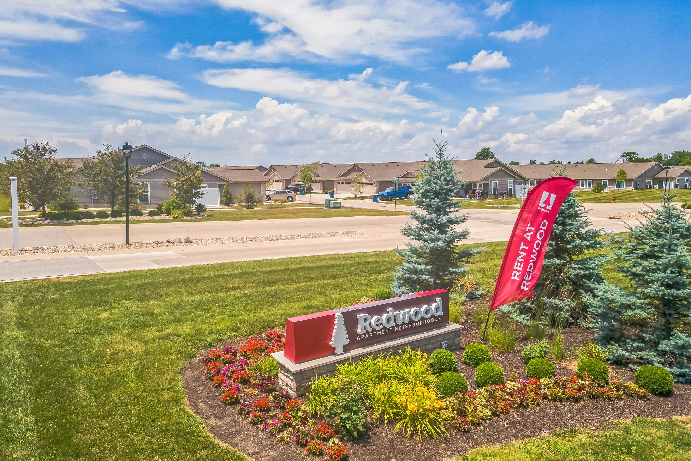 Entrance sign for Redwood Apartments & Townhomes, surrounded by landscaped flowers and bushes, with a red "Rent at Redwood" flag and a suburban neighborhood in the background.