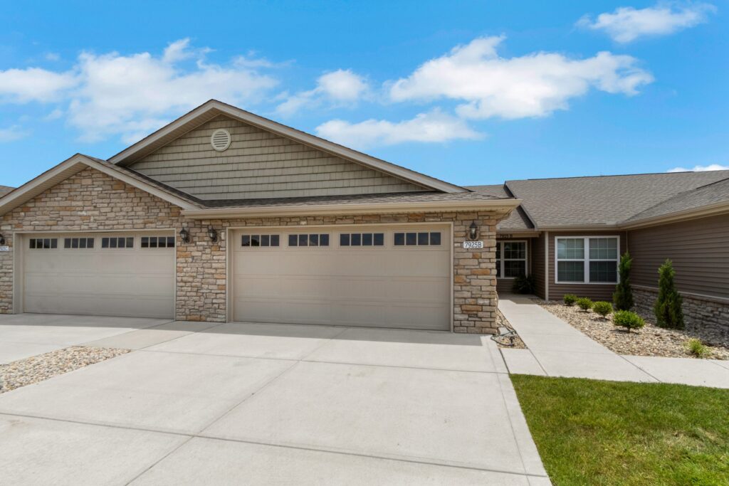 Single-story house with stone and siding exterior, three-car garage, paved driveway, and minimal landscaping under a partly cloudy sky.