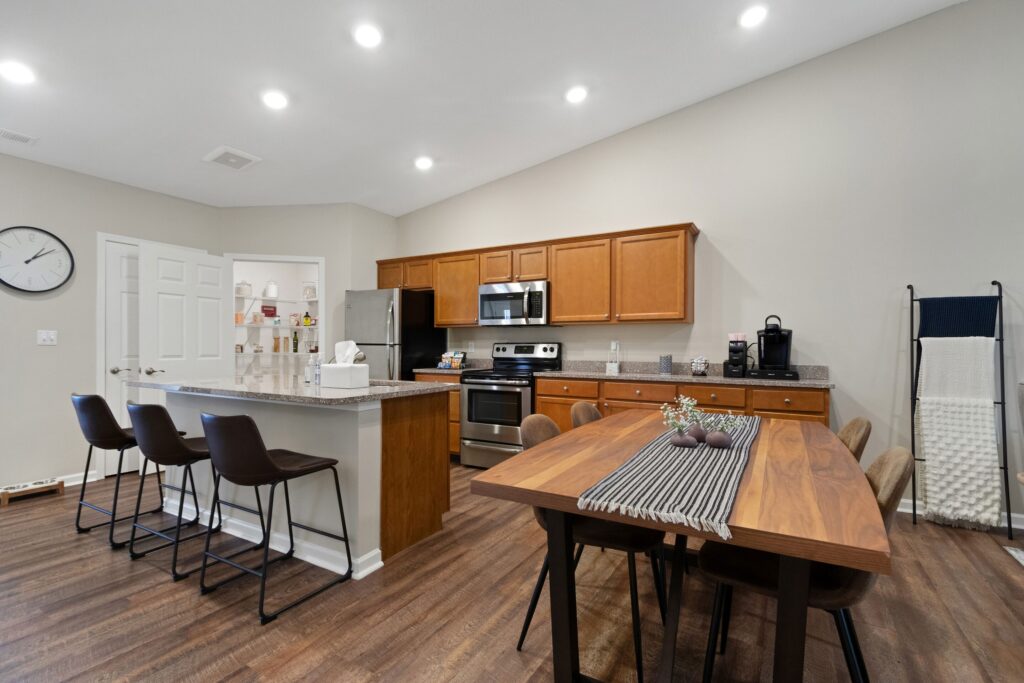 Modern kitchen and dining area with wooden cabinets, stainless steel appliances, island with three barstools, and a wooden table set for six on hardwood floors under recessed lighting.
