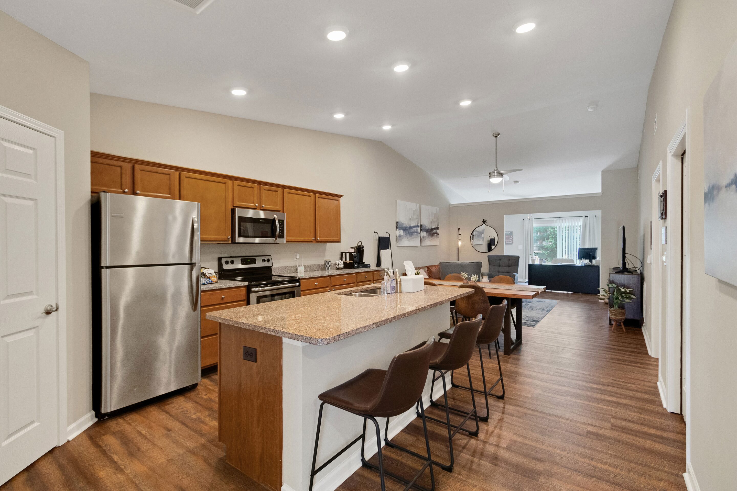 Modern kitchen with stainless steel appliances, wooden cabinets, and an island with three barstools, opening to a living area with a sofa and large window in the background.