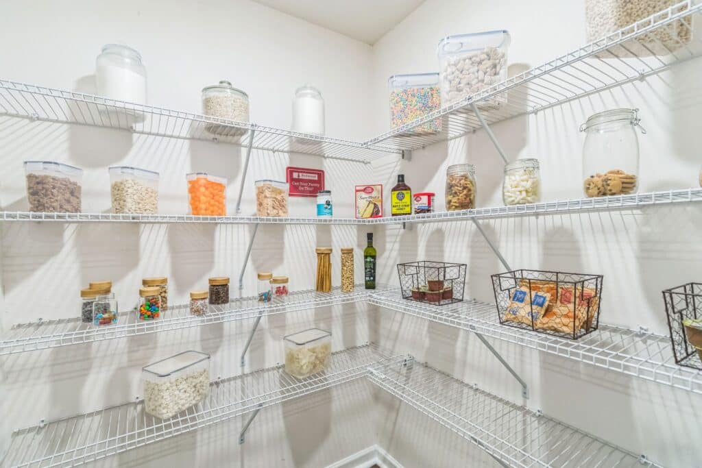 Organized pantry with wire shelves holding clear containers of cereals, snacks, and dry goods, along with jars, bottles, and wire baskets storing various food items.