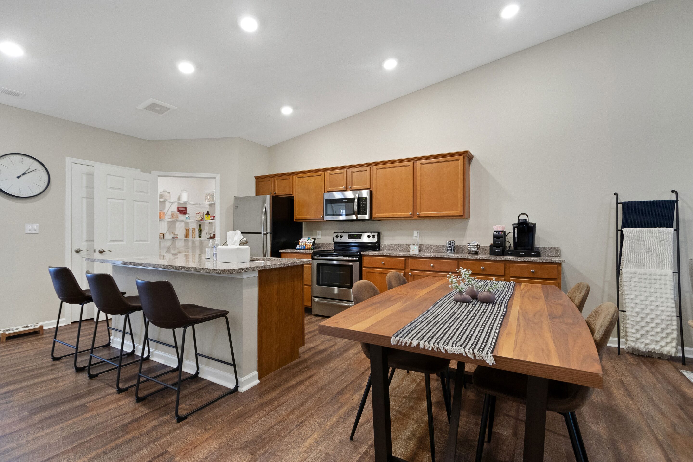 Modern kitchen and dining area with wooden cabinets, stainless steel appliances, island with three barstools, and a wooden table set for six on hardwood floors under recessed lighting.