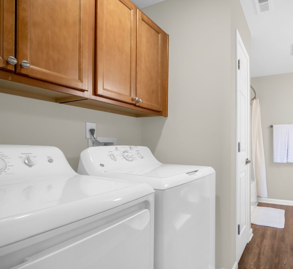 A laundry room with a white washing machine and dryer, wooden cabinets above, beige walls, and a view into a bathroom with a towel on a rack.