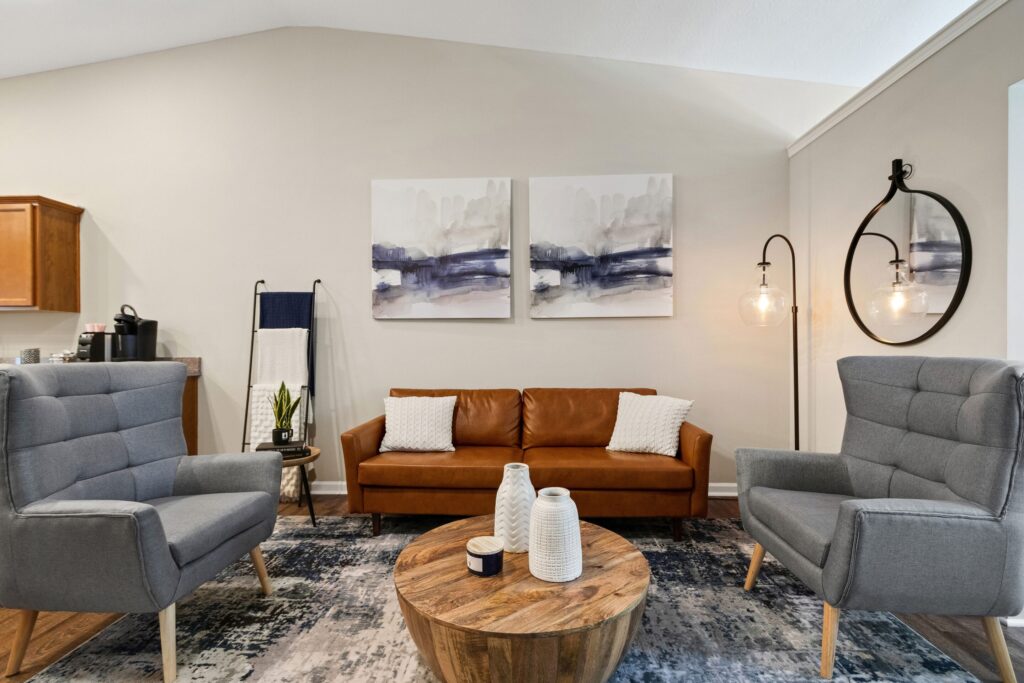 Modern living room with a brown leather sofa, two gray armchairs, a round wooden coffee table, abstract wall art, and a hanging light fixture with a round mirror.