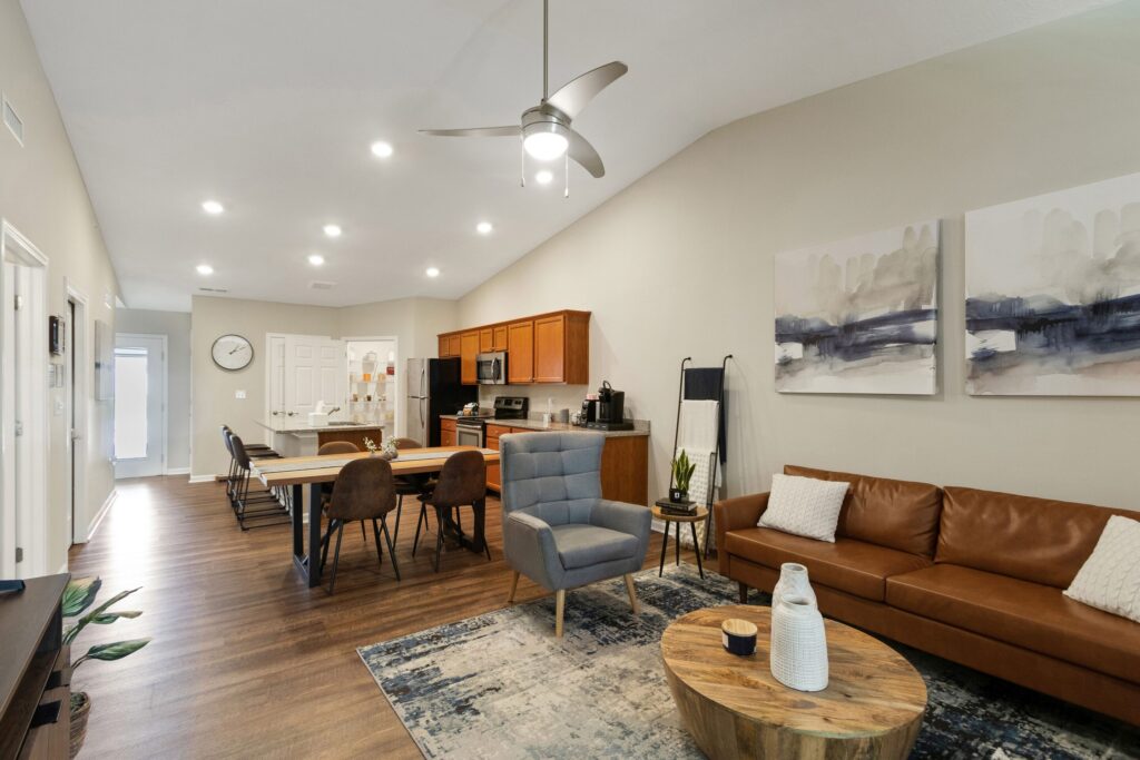 Open-concept living and dining area with brown leather sofa, modern armchair, round wooden coffee table, dining table, kitchen, ceiling fan, and abstract wall art.