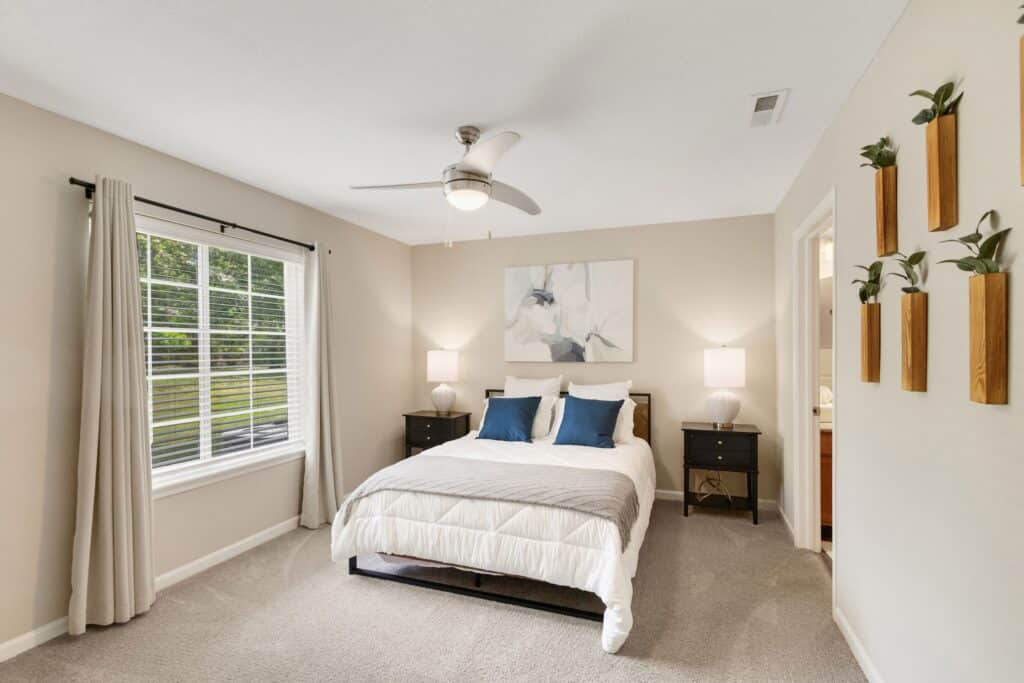 A neatly made bed with white bedding and blue pillows is centered between two nightstands in a bright, neutral bedroom with a window, wall art, and potted plants.