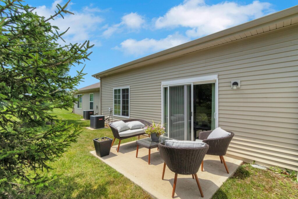 Outdoor patio with wicker chairs, bench seating, and a small table on a concrete slab beside a beige house with sliding glass doors and grassy yard.