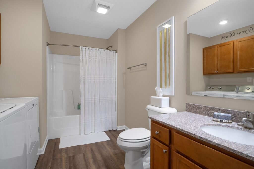 Bathroom with a shower-tub combo, white curtain, toilet, sink with granite countertop, wooden cabinets, and a washer and dryer unit on the left side.