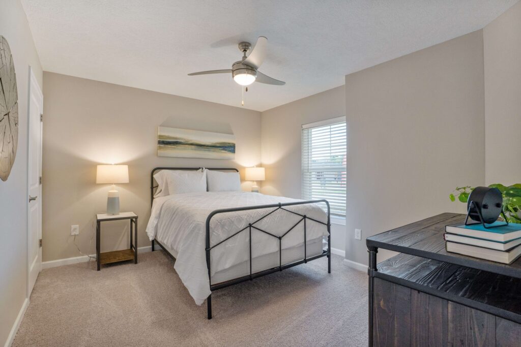 A bedroom with a metal-frame bed, white bedding, two bedside tables with lamps, a ceiling fan, a dresser with books and a plant, and a window with blinds.