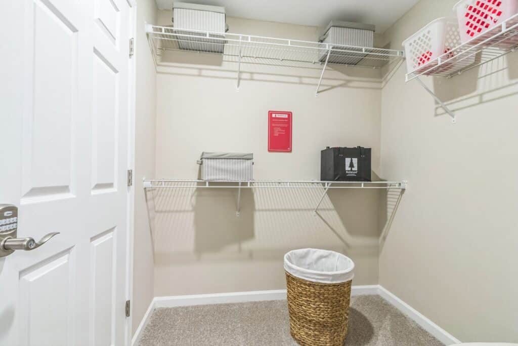 A laundry room with wire shelves, storage boxes, laundry baskets, a red sign on the wall, and a wicker laundry hamper on the carpeted floor.